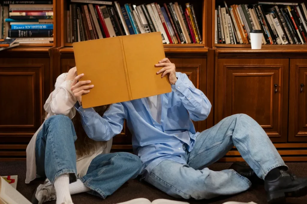 Duas pessoas sentadas no chão de uma biblioteca, escondendo o rosto atrás de um livro aberto