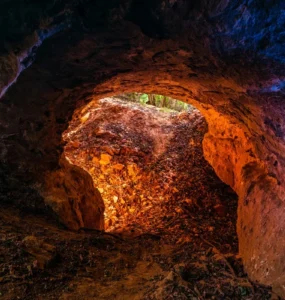 Entrada de uma caverna com paredes rochosas iluminadas em tons alaranjados e azulados, mostrando folhas secas no chão e a saída para a natureza ao fundo.