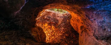 Entrada de uma caverna com paredes rochosas iluminadas em tons alaranjados e azulados, mostrando folhas secas no chão e a saída para a natureza ao fundo.