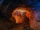 Entrada de uma caverna com paredes rochosas iluminadas em tons alaranjados e azulados, mostrando folhas secas no chão e a saída para a natureza ao fundo.