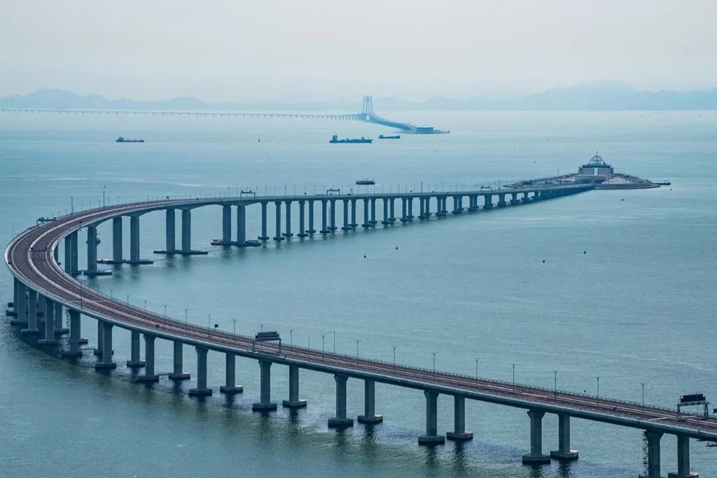 Vista panorâmica da Ponte Hong Kong–Zhuhai–Macau atravessando o mar, com curvas elegantes e pilares altos sobre águas calmas, ao fundo montanhas e embarcações.