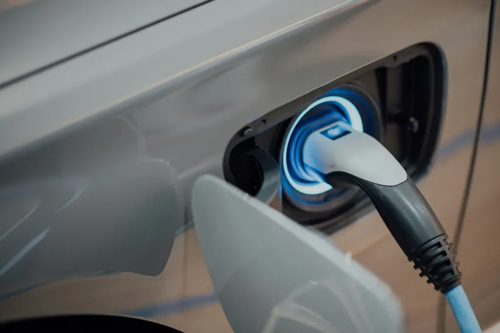 Close-up de carregador conectado à entrada de energia de um carro elétrico prateado, com luz azul indicando processo de recarga em andamento
