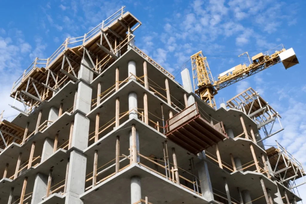 Estrutura de concreto armado de um edifício em construção com grua amarela ao fundo e céu azul com nuvens claras.