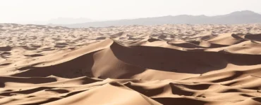 Vista ampla de dunas do deserto em tons dourados, iluminadas pelo sol com montanhas ao fundo.