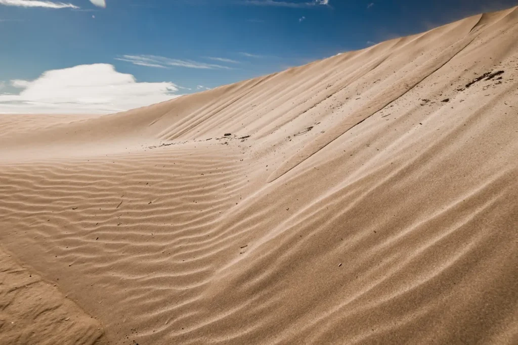 Duna de areia inclinada com marcas onduladas do vento sob céu azul com nuvens brancas.