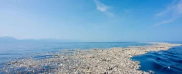 Imagem de uma imensa mancha de poluição plástica no oceano, conhecida como ilha de lixo, mostrando garrafas, embalagens e detritos acumulados na superfície da água sob um céu azul.