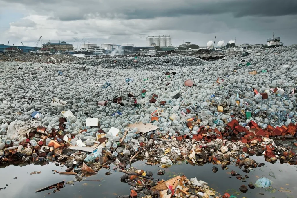 Imagem de um enorme acúmulo de garrafas plásticas, embalagens e resíduos em um lixão a céu aberto próximo a uma área urbana com prédios e barcos ao fundo.