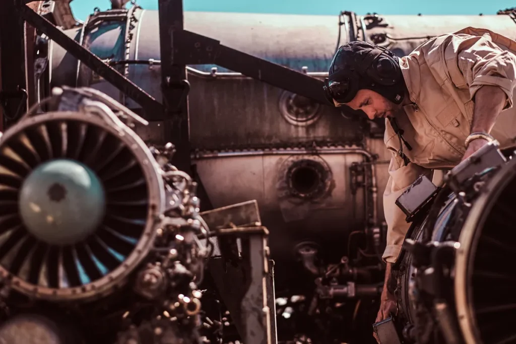Engenheiro aeronáutico com uniforme bege e capacete de proteção inspeciona turbina a jato em ambiente industrial.