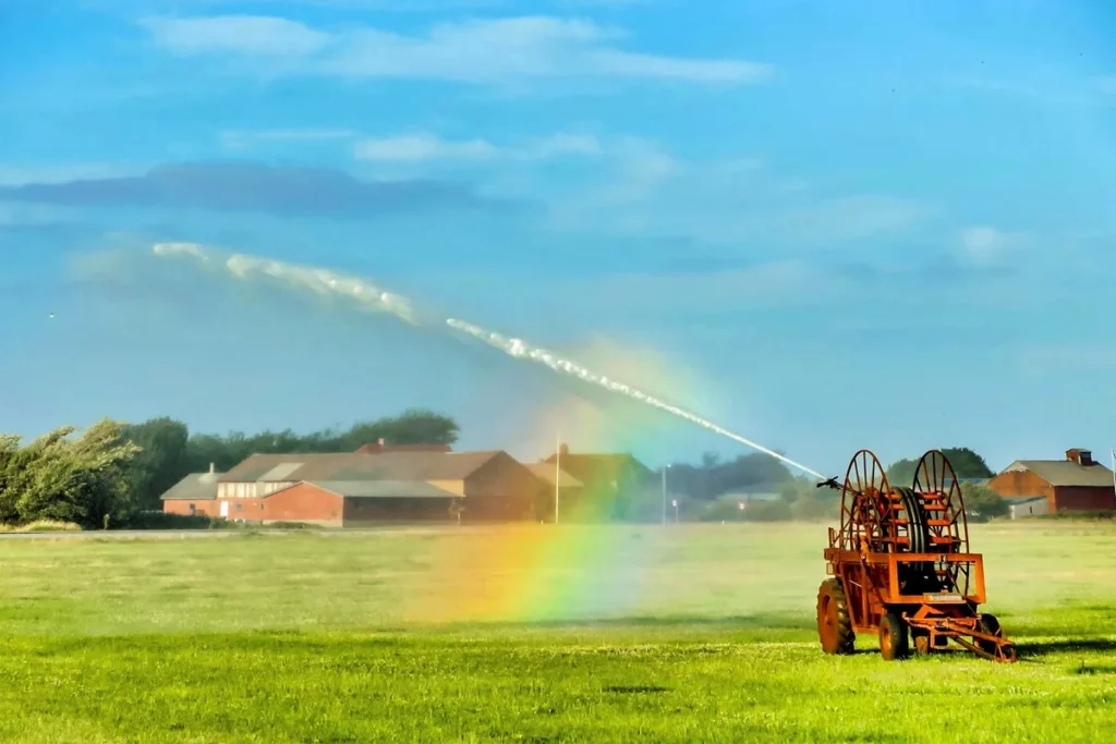 Equipamento de irrigação agrícola lança jato de água em campo verde, formando um arco-íris sob o céu azul.