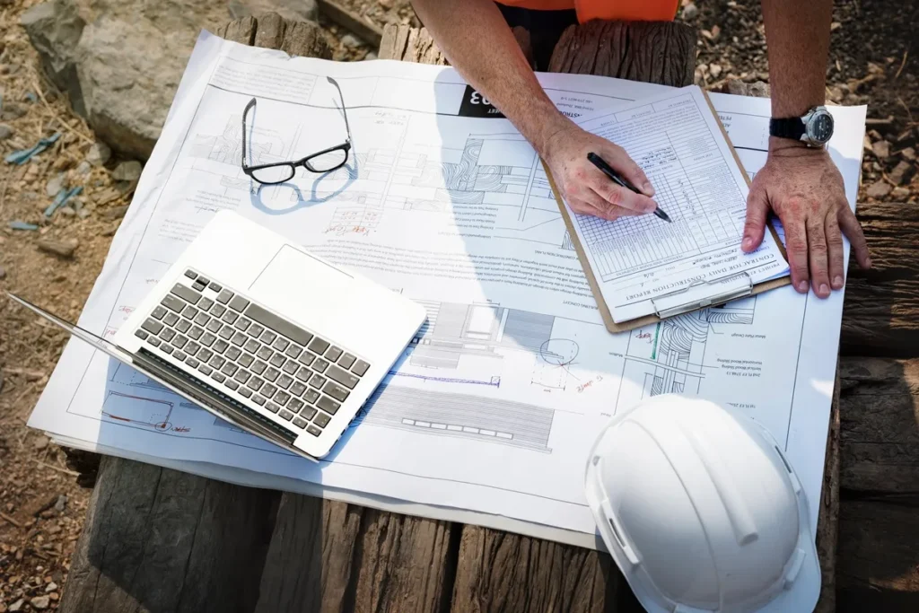 Engenheiro analisa documentos de topografia e planejamento de obra em prancheta, com notebook, óculos e capacete de segurança sobre a mesa.