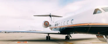 Jato executivo branco e preto estacionado em uma pista de aeroporto, com céu parcialmente nublado e tons suaves de azul e branco ao fundo.