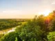Imagem de uma agrofloresta banhada pela luz do pôr do sol, com árvores verdes, um rio serpenteando ao centro e o céu dourado ao fundo.