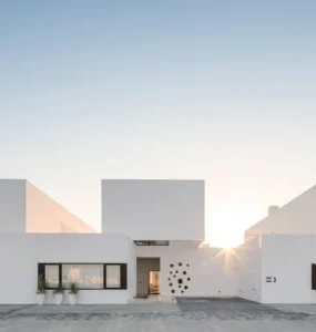 Fachada de casas com arquitetura minimalista e design moderno, com volumes geométricos brancos e linhas retas sob um céu azul claro ao pôr do sol.