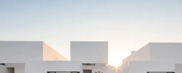 Fachada de casas com arquitetura minimalista e design moderno, com volumes geométricos brancos e linhas retas sob um céu azul claro ao pôr do sol.