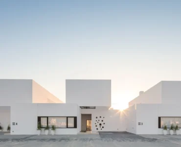 Fachada de casas com arquitetura minimalista e design moderno, com volumes geométricos brancos e linhas retas sob um céu azul claro ao pôr do sol.