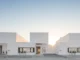 Fachada de casas com arquitetura minimalista e design moderno, com volumes geométricos brancos e linhas retas sob um céu azul claro ao pôr do sol.
