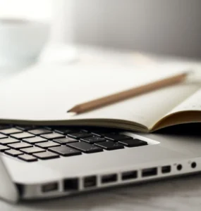 Notebook prateado aberto com teclado preto, caderno aberto e lápis apoiado sobre as páginas, em uma mesa clara com uma xícara desfocada ao fundo.