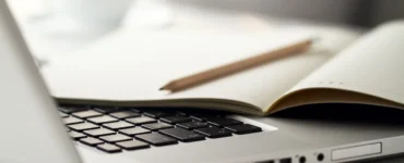 Notebook prateado aberto com teclado preto, caderno aberto e lápis apoiado sobre as páginas, em uma mesa clara com uma xícara desfocada ao fundo.