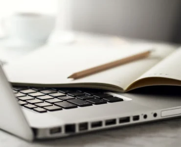 Notebook prateado aberto com teclado preto, caderno aberto e lápis apoiado sobre as páginas, em uma mesa clara com uma xícara desfocada ao fundo.