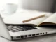 Notebook prateado aberto com teclado preto, caderno aberto e lápis apoiado sobre as páginas, em uma mesa clara com uma xícara desfocada ao fundo.