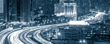Vista noturna de uma cidade inteligente com arranha-céus iluminados e rodovias elevadas cheias de luzes de tráfego em movimento.