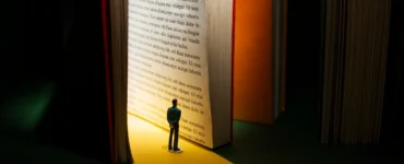 Miniatura de pessoa observando um livro gigante iluminado, representando a busca pelo conhecimento e a importância da divulgação científica.
