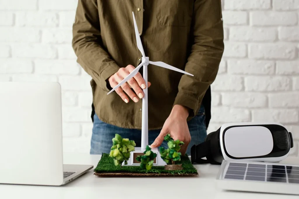 Pessoa ajustando maquete de turbina eólica em mesa com mini árvores, laptop e painel solar, simbolizando inovação e eficiência energética sustentável.