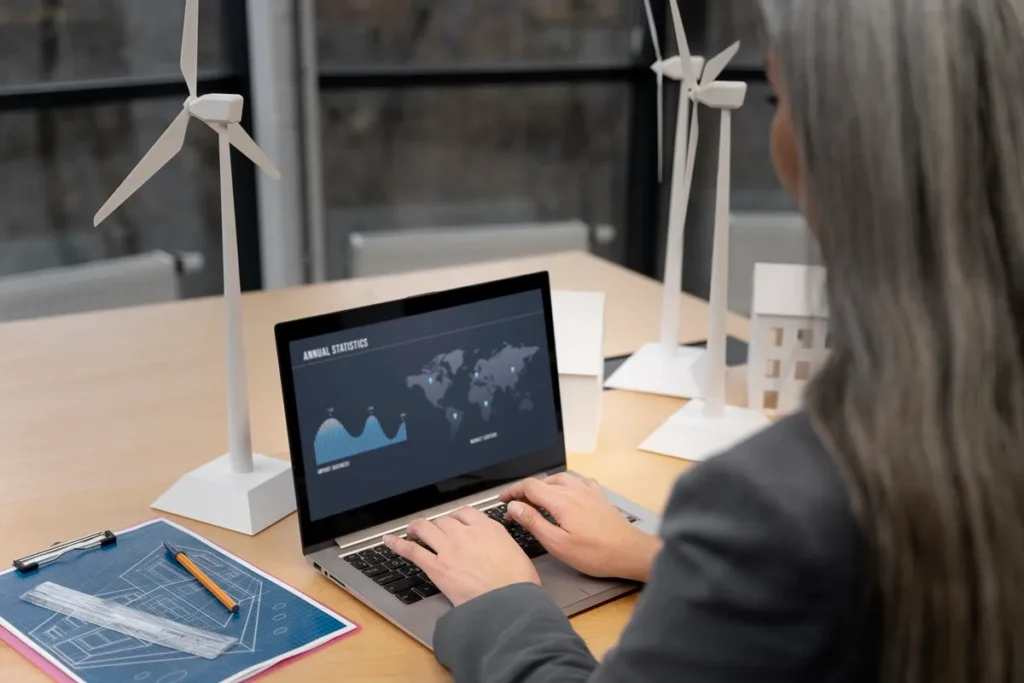Profissional analisando gráficos de eficiência energética em laptop, ao lado de maquetes de turbinas eólicas e projetos de arquitetura em mesa de escritório.