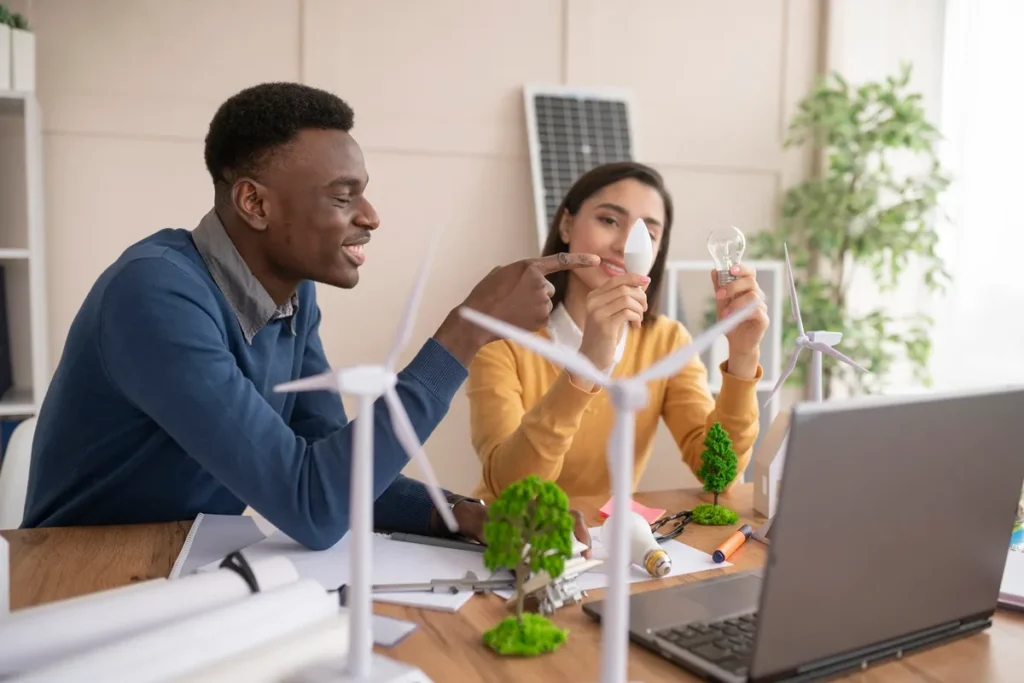Dois jovens engenheiros discutindo ideias sobre energia renovável, com maquetes de turbinas eólicas e painéis solares sobre a mesa.