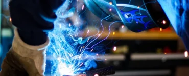 Soldador com máscara de proteção e luvas realizando soldagem elétrica com faíscas azuis em ambiente industrial.