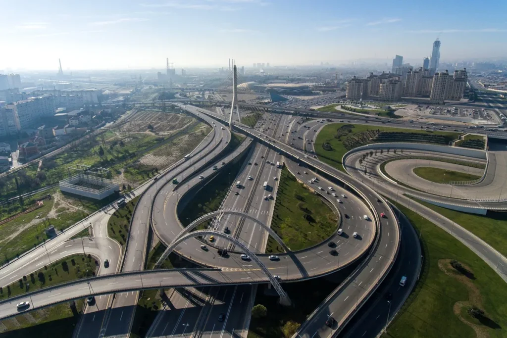 Vista aérea de um complexo viário moderno durante o dia, com múltiplas pistas, curvas e viadutos rodeados por áreas verdes, representando a engenharia de transportes e o planejamento urbano.