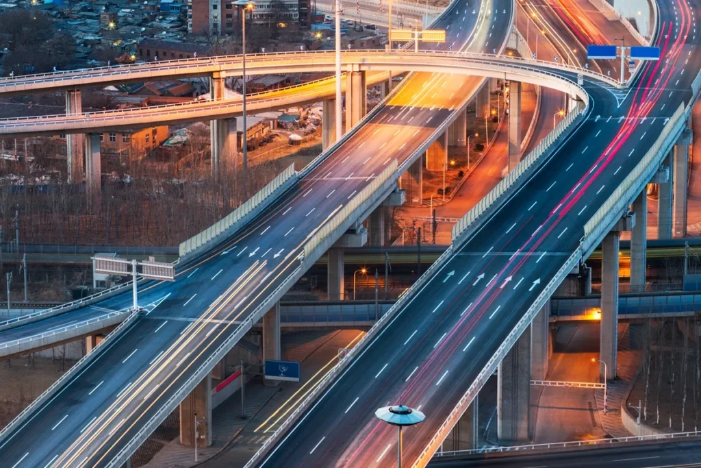 Imagem de rodovias elevadas e viadutos iluminados ao entardecer, com várias pistas largas e fluxo de veículos, representando a engenharia de transportes e a infraestrutura urbana moderna