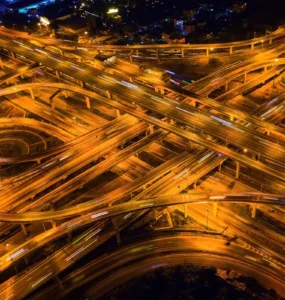 Vista aérea de um complexo viário iluminado à noite, mostrando várias pistas elevadas e curvas que se cruzam, representando a engenharia de transportes e a mobilidade urbana moderna.