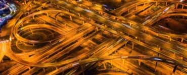 Vista aérea de um complexo viário iluminado à noite, mostrando várias pistas elevadas e curvas que se cruzam, representando a engenharia de transportes e a mobilidade urbana moderna.