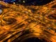 Vista aérea de um complexo viário iluminado à noite, mostrando várias pistas elevadas e curvas que se cruzam, representando a engenharia de transportes e a mobilidade urbana moderna.