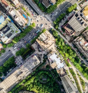 Imagem aérea de uma cidade com ruas largas, cruzamentos movimentados, prédios altos e árvores distribuídas entre as construções, representando a organização e o crescimento urbano.