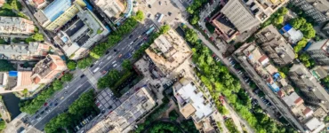 Imagem aérea de uma cidade com ruas largas, cruzamentos movimentados, prédios altos e árvores distribuídas entre as construções, representando a organização e o crescimento urbano.