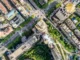 Imagem aérea de uma cidade com ruas largas, cruzamentos movimentados, prédios altos e árvores distribuídas entre as construções, representando a organização e o crescimento urbano.
