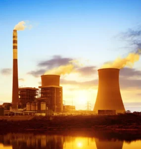Usina termelétrica com torres de resfriamento e chaminé liberando vapor sob o pôr do sol, refletida em um lago, representando a geração de energia térmica.