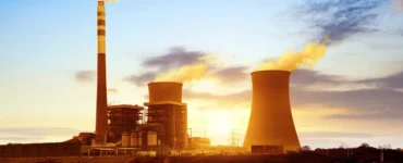 Usina termelétrica com torres de resfriamento e chaminé liberando vapor sob o pôr do sol, refletida em um lago, representando a geração de energia térmica.