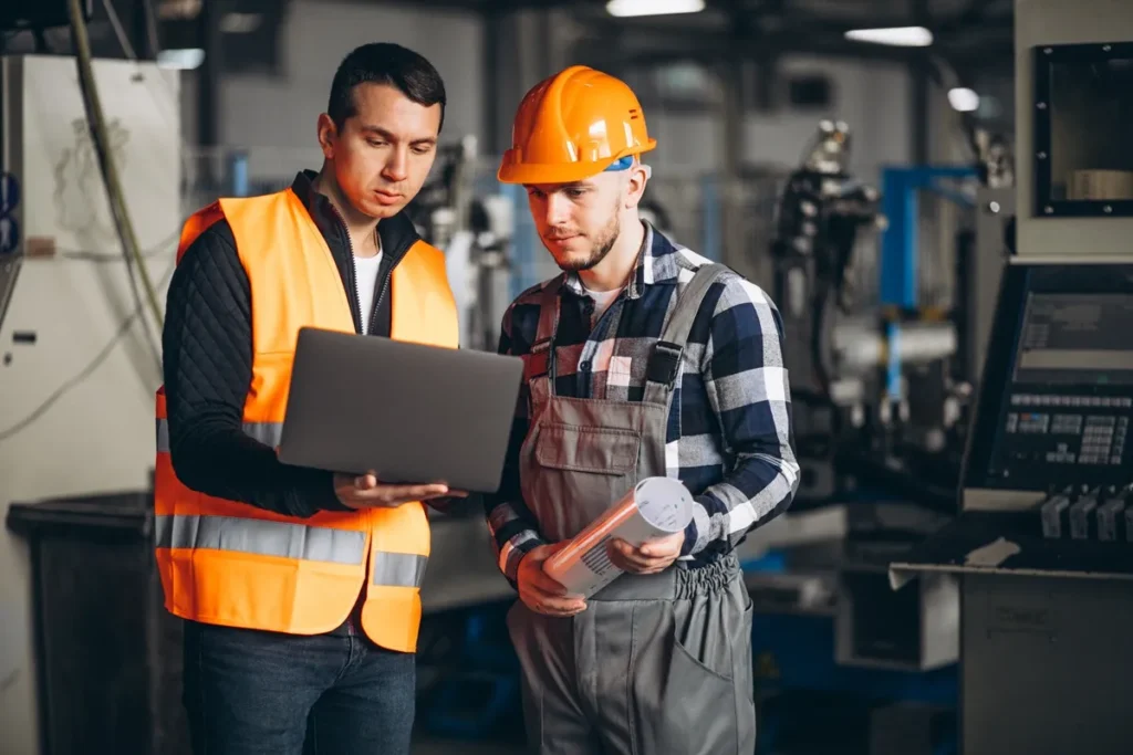 Dois engenheiros industriais, um com colete refletivo e outro com capacete laranja, analisando dados em um laptop dentro de uma fábrica moderna.