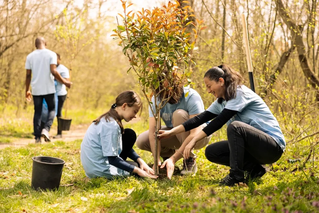 Grupo de voluntários sorridentes, incluindo uma criança e dois adultos, plantando uma muda de árvore em um parque durante uma ação de reflorestamento e preservação ambiental.