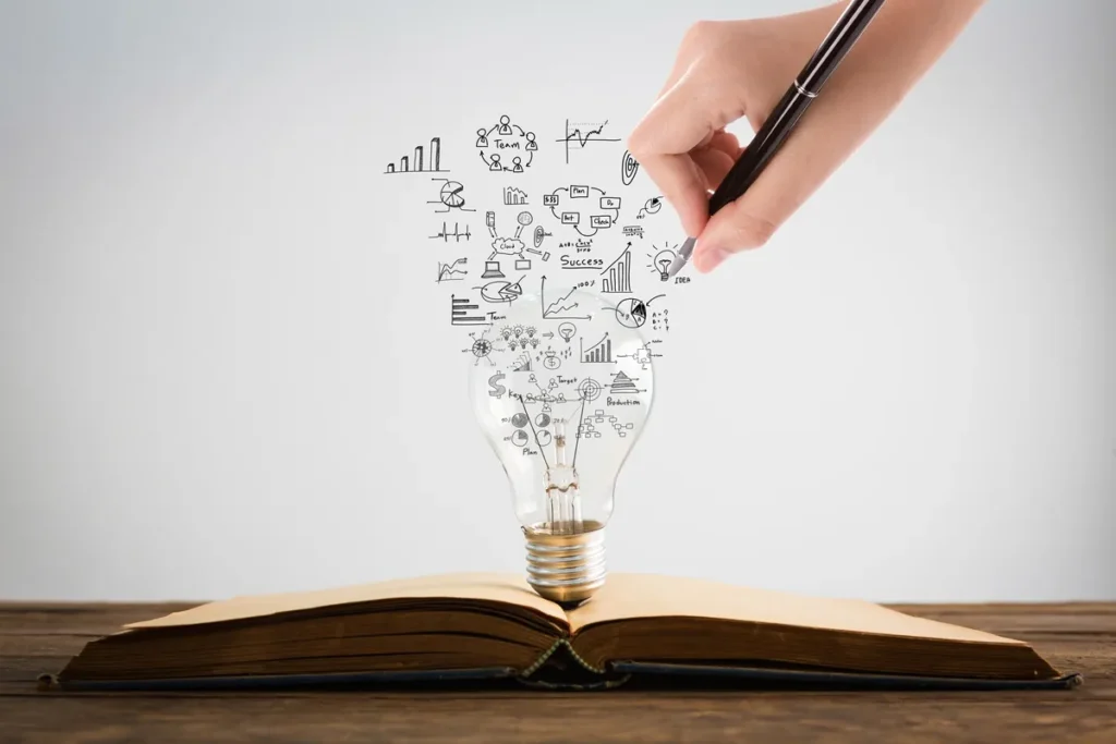 Mão segurando uma caneta desenhando ideias e gráficos sobre uma lâmpada posicionada em cima de um livro aberto, simbolizando criatividade e conhecimento científico.