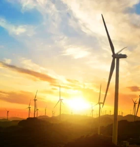 Parque eólico com várias turbinas de vento em funcionamento ao pôr do sol, com céu colorido em tons de laranja e azul, simbolizando energia renovável e sustentabilidade.