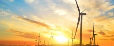 Parque eólico com várias turbinas de vento em funcionamento ao pôr do sol, com céu colorido em tons de laranja e azul, simbolizando energia renovável e sustentabilidade.