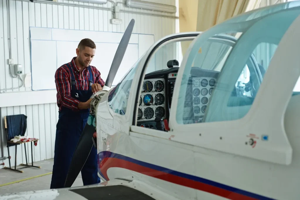 Mecânico de aviação inspecionando o motor e a hélice de uma aeronave leve dentro de um hangar.
