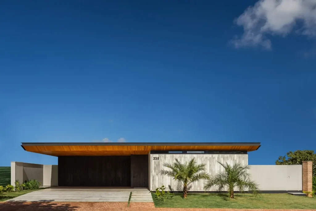 Fachada de casa térrea moderna e minimalista com linhas retas, cobertura em balanço com forro de madeira e paisagismo tropical com palmeiras sob céu azul.