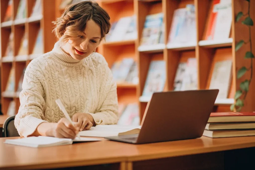 Mulher usando suéter de tricô claro estuda em uma biblioteca, escrevendo em um caderno com um notebook aberto e pilhas de livros sobre a mesa.