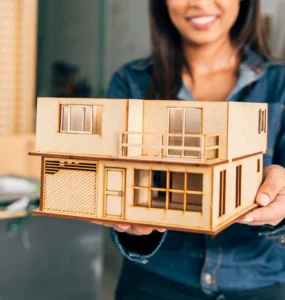 Mulher sorridente segura uma maquete arquitetônica de casa moderna de dois andares feita em madeira, com janelas, varanda e garagem em miniatura.