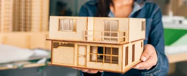 Mulher sorridente segura uma maquete arquitetônica de casa moderna de dois andares feita em madeira, com janelas, varanda e garagem em miniatura.